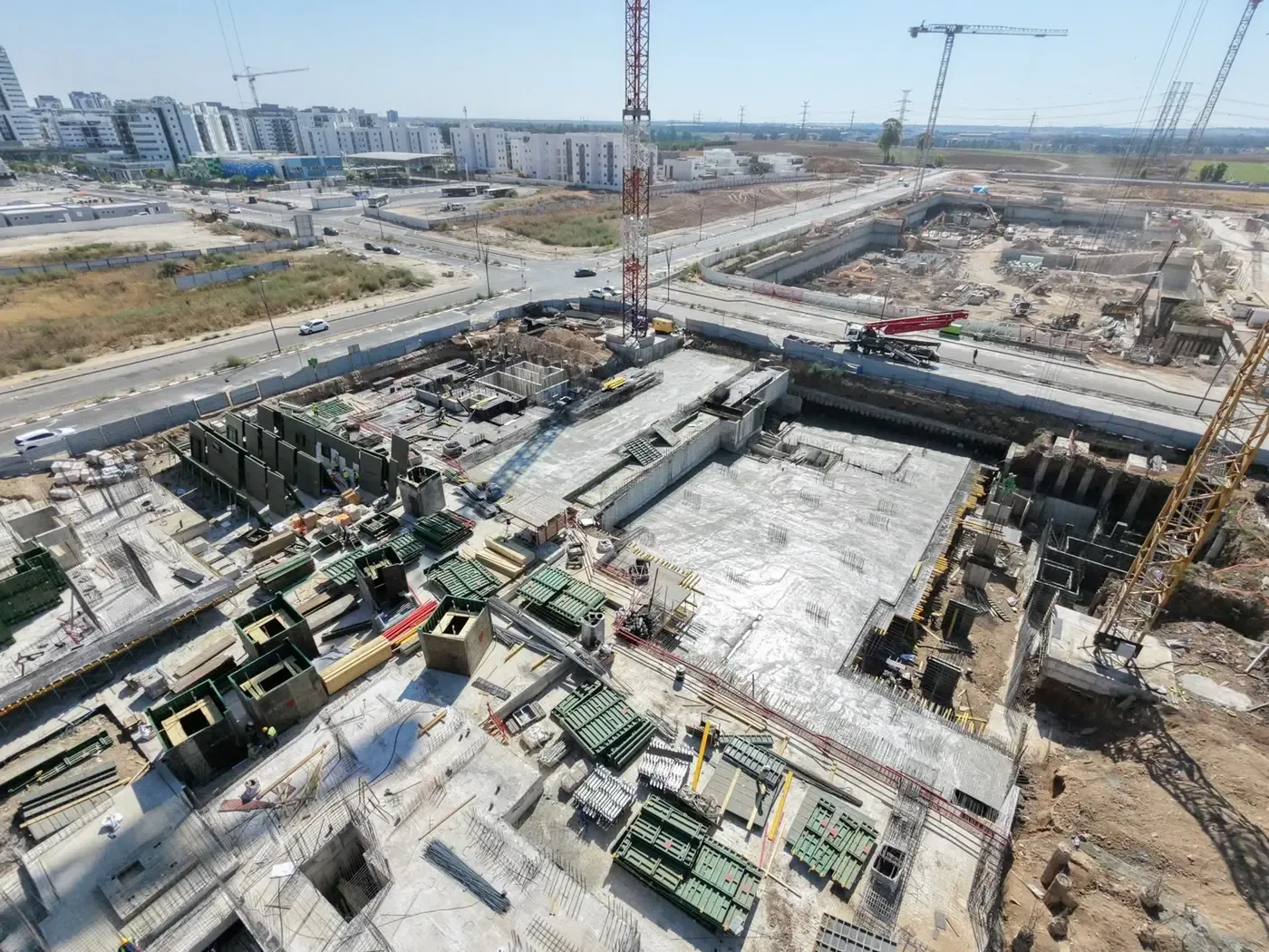 צילום אוויר של אתר הבנייה בכרמי גת: תקרת מרתף יצוקה, טפסות קירות ועגורני צריח, ומאחור השכונות הבנויות של קרית גת
