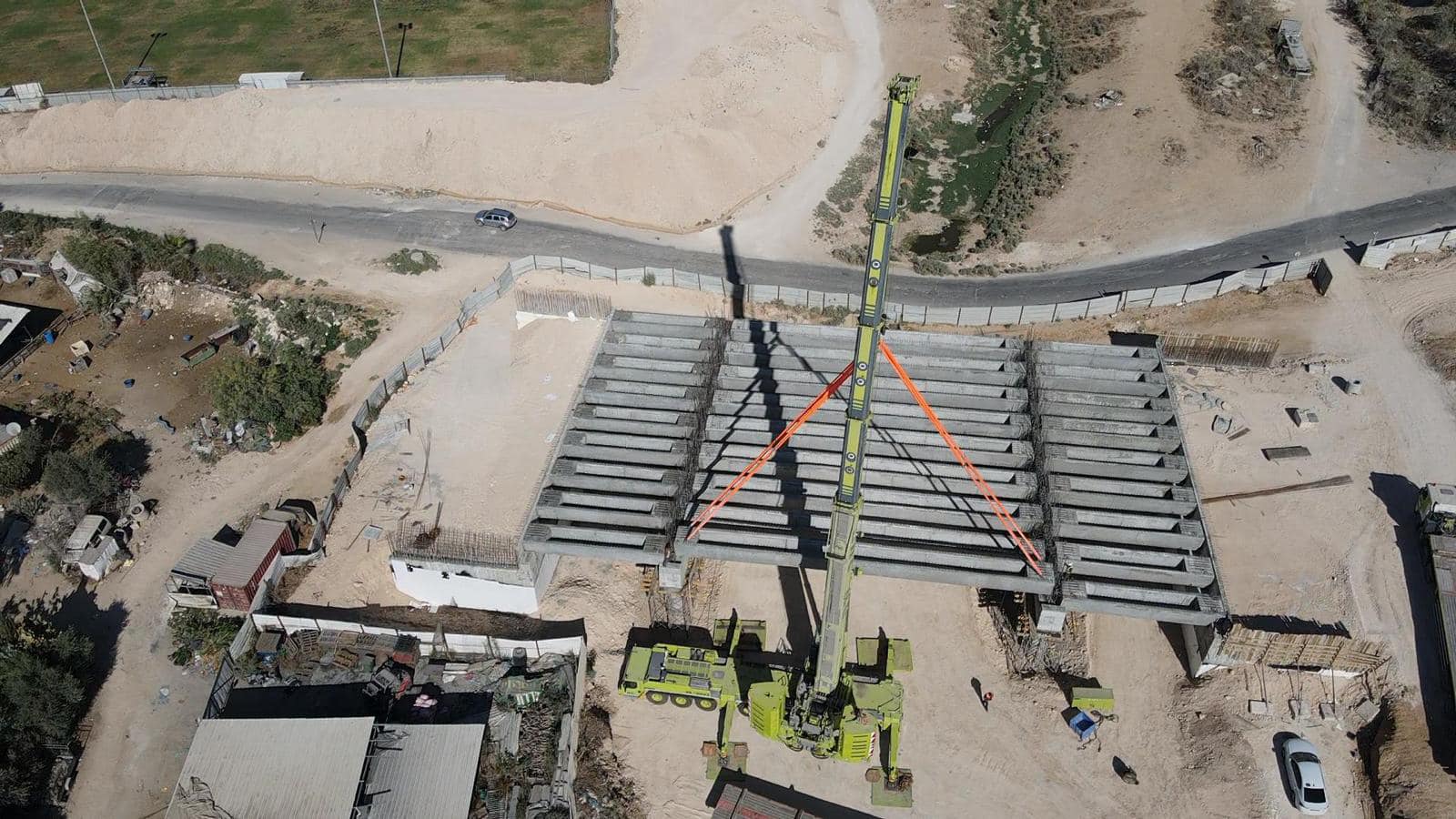 מנוף צהוב גדול מניח קורות בטון טרומיות על ראשי העמודים של הגשר, במבט מלמעלה