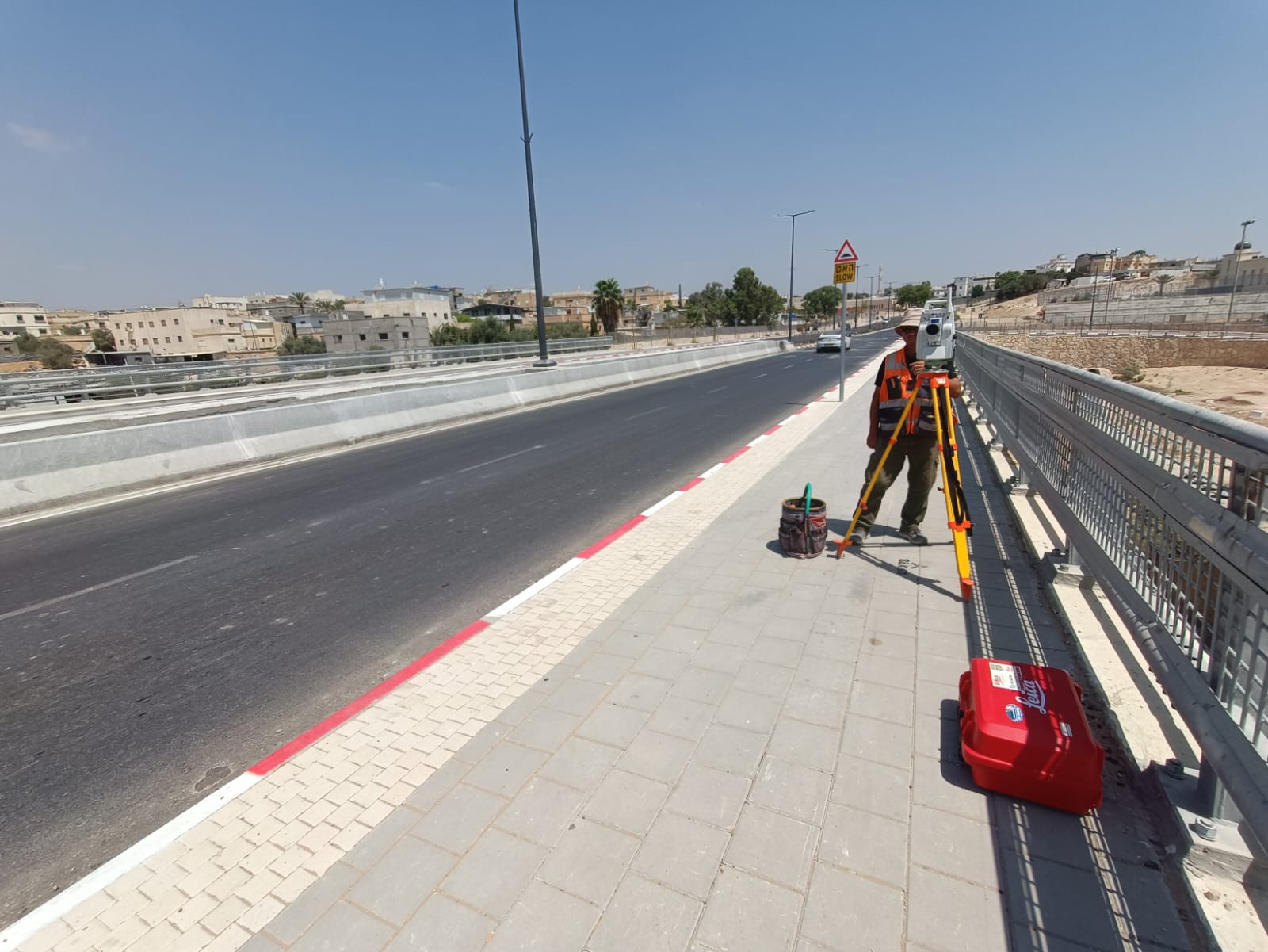 מודד מהצוות שלנו עומד על מדרכת הגשר עם תחנה טוטאלית לייקה על חצובה, מודד את הכביש הגמור