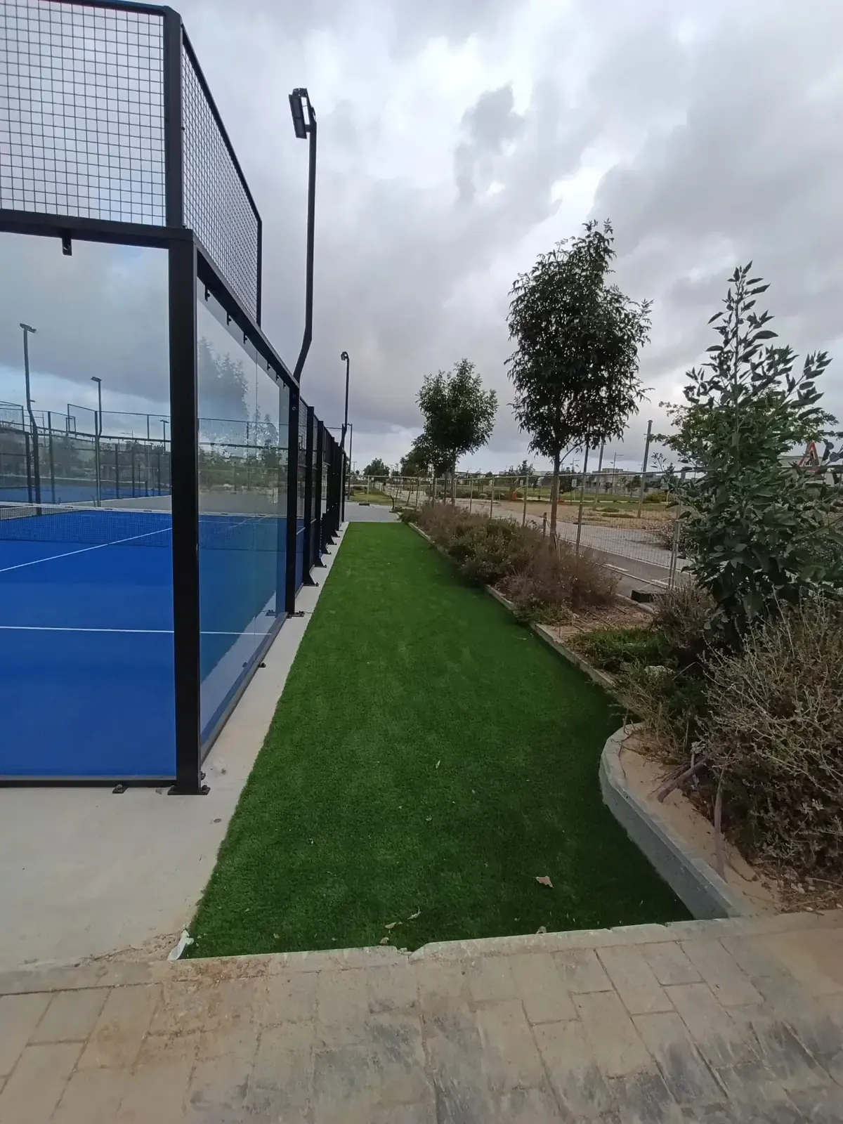 רצועת דשא סינתטי וערוגות גינון לאורך דופן הזכוכית של מגרש הפאדל