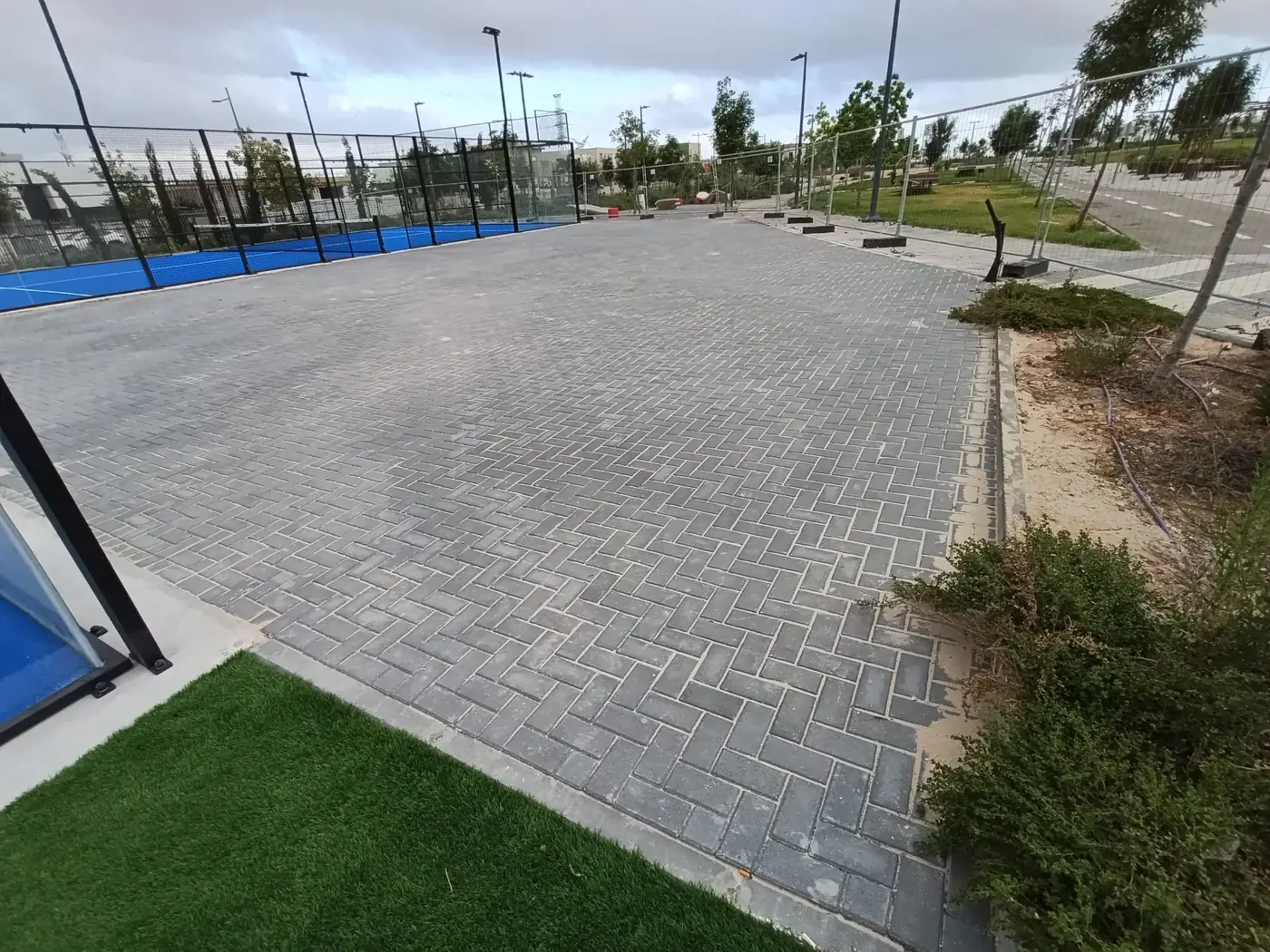 משטח ריצוף רחב בין מגרשי הפאדל לגדר המתחם, עם עמודי תאורה ושכונת שרונית ברקע