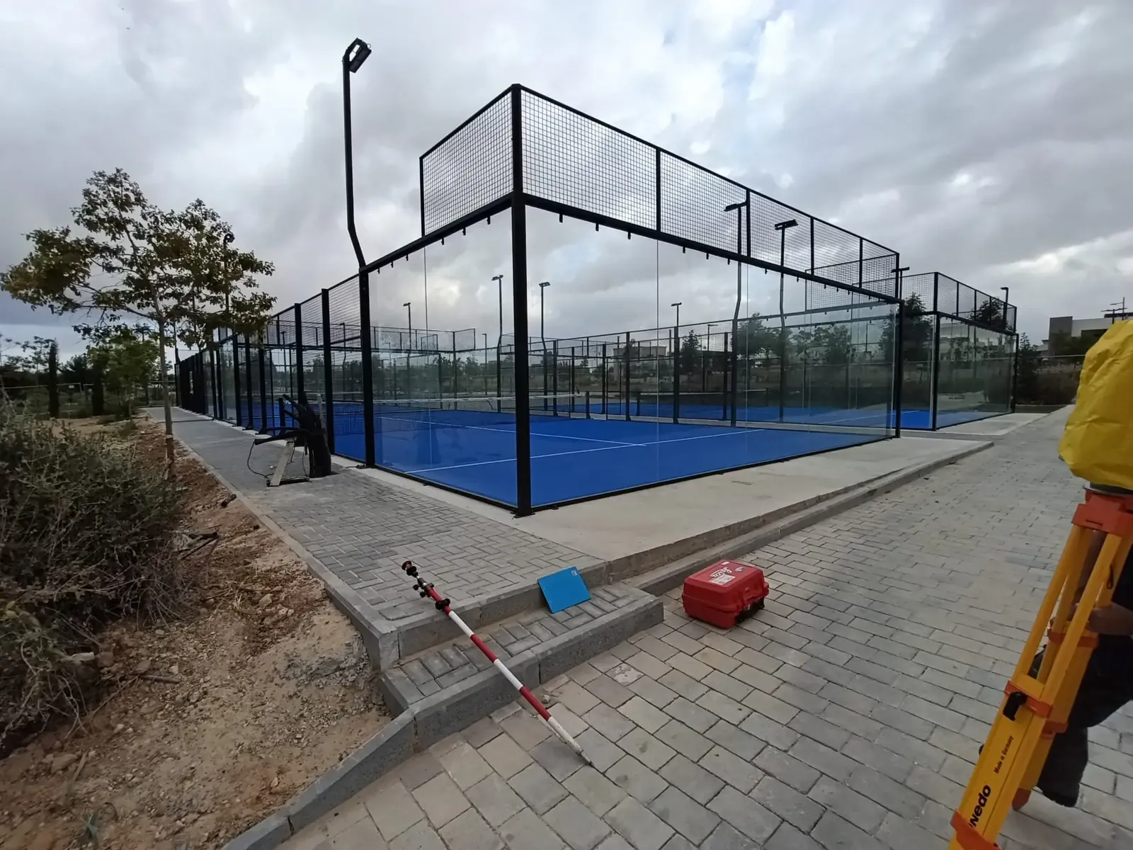 חצובה, ארגז מכשור ומוט פריזמה מונחים על הריצוף מול מגרשי הפאדל בלהבים